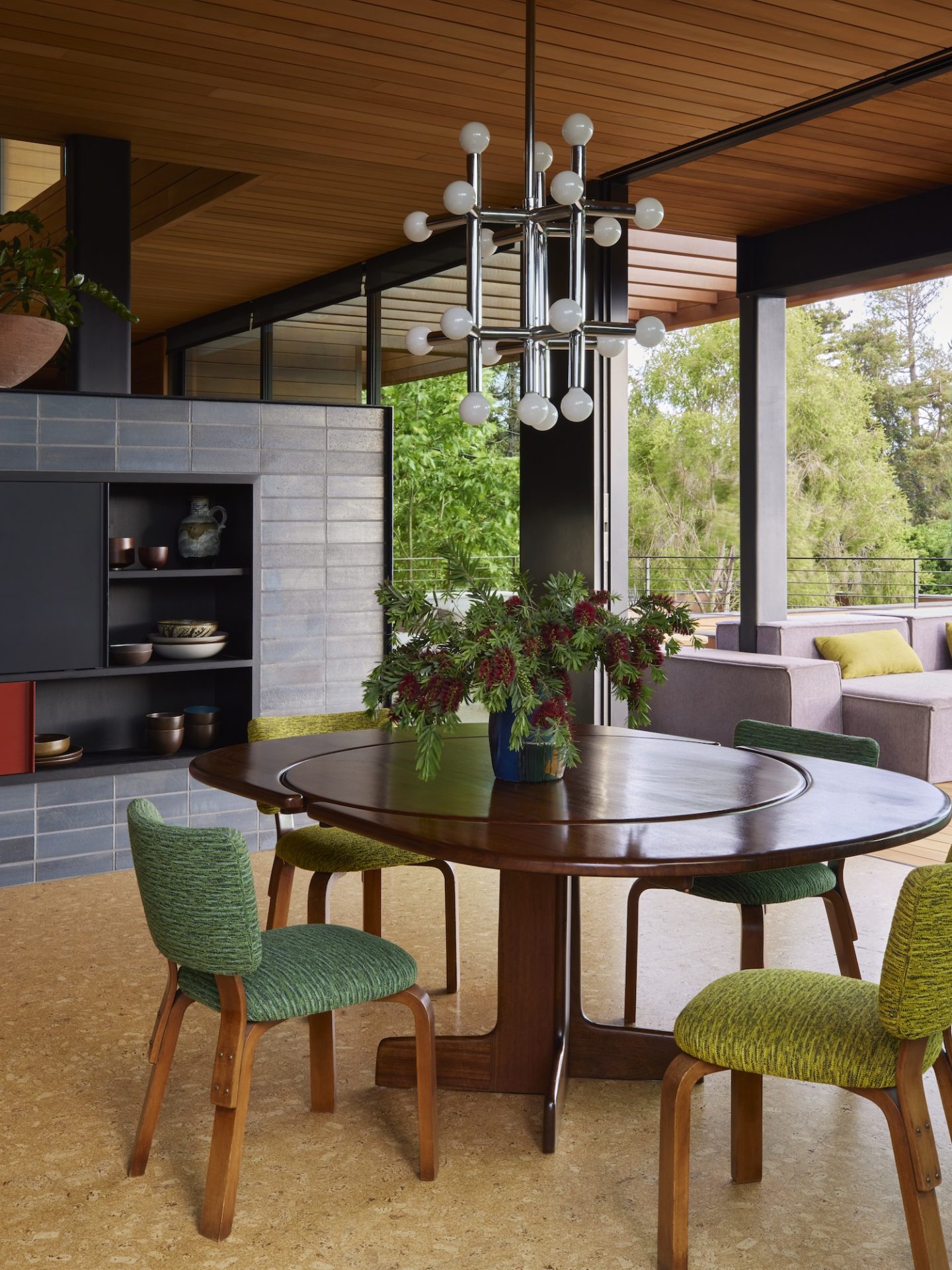 A dining area with a round table and green chairs. A chrome chandelier hangs above, highlighting the 70s interior design and open layout.