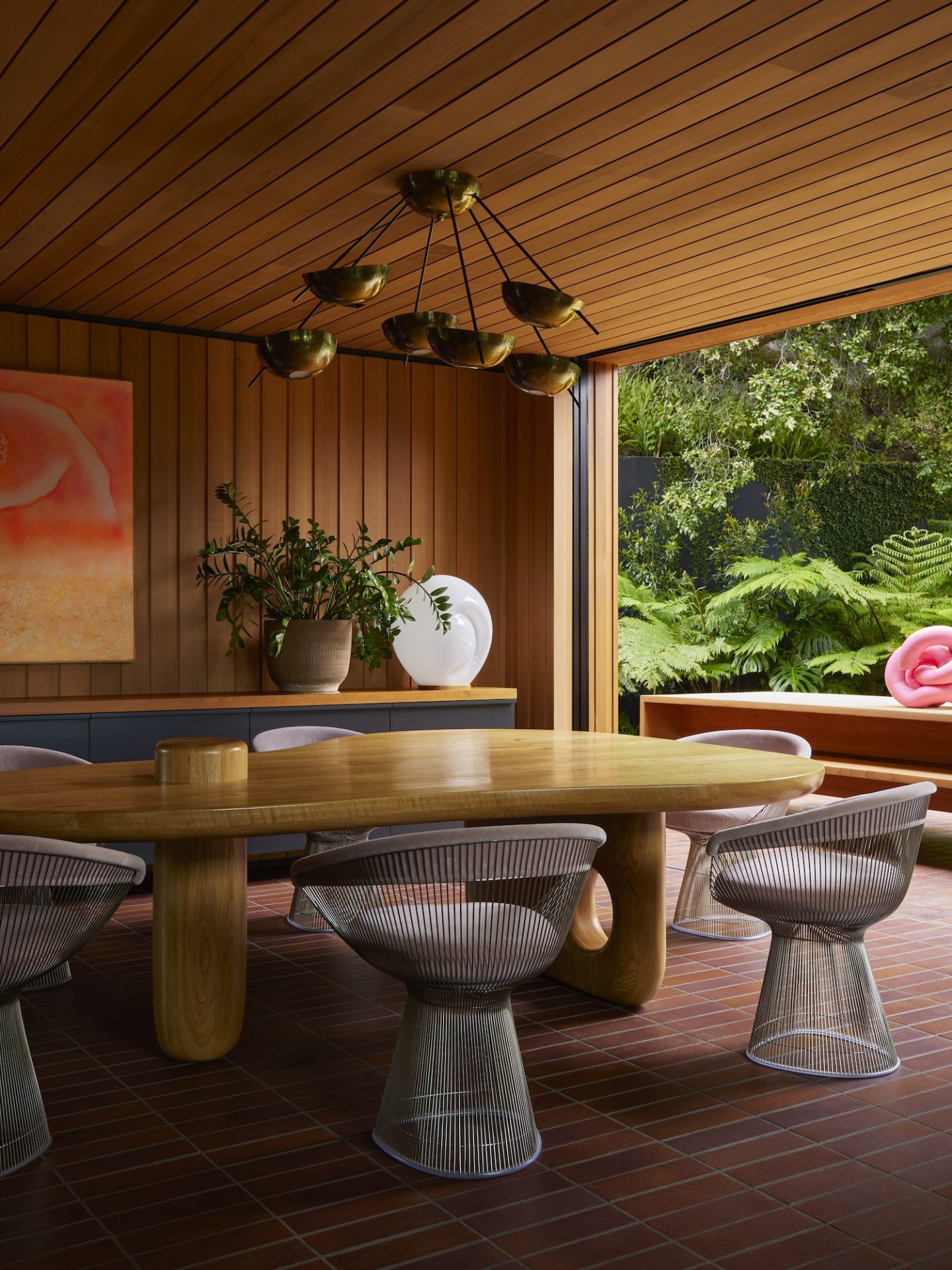 A dining room with a long wooden table and unique wire chairs. Warm wood paneling and a brass chandelier evoke 70s interior design charm.