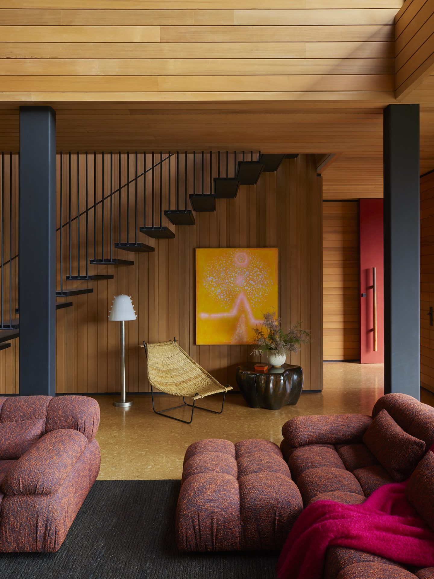 A living space with a staircase and warm wood paneling. Plush sofas and a vibrant painting enhance the 70s interior design vibe.