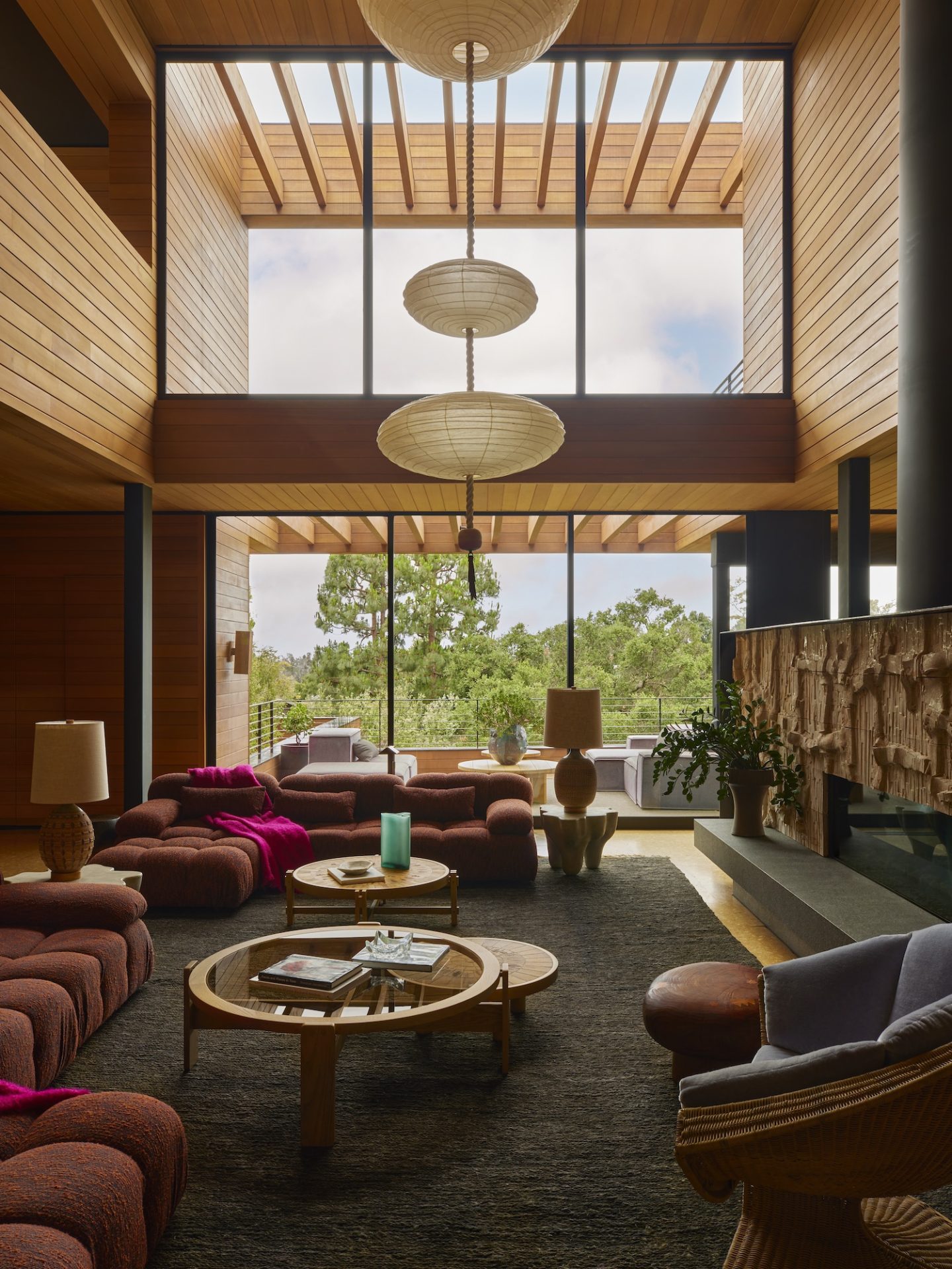 A double-height living room with warm wood ceilings and large windows. Plush seating and unique light fixtures capture the 70s interior design feel.