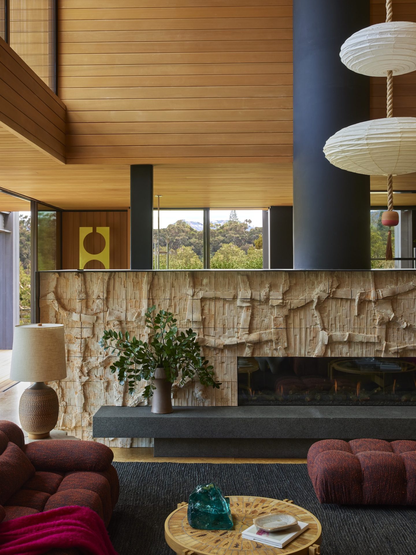 A living room with a textured stone fireplace and warm wood ceilings. Pendant lights and plush seating evoke a cozy 70s interior design atmosphere.