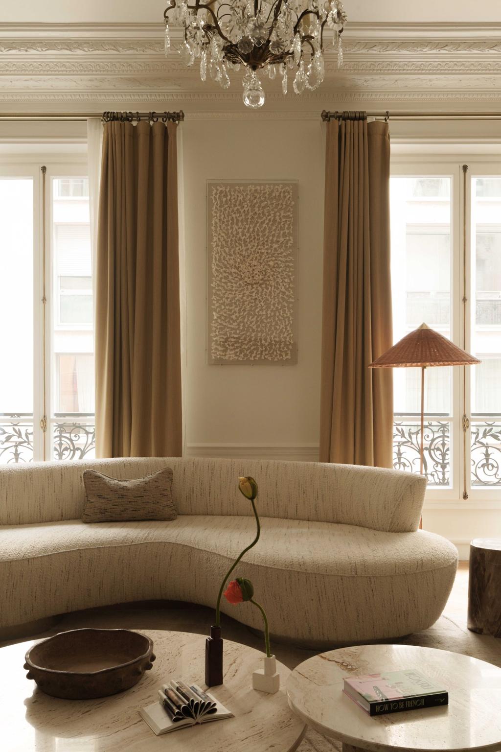 Curved cream sofa by tall windows with marble coffee table in a light-filled Parisian living room.