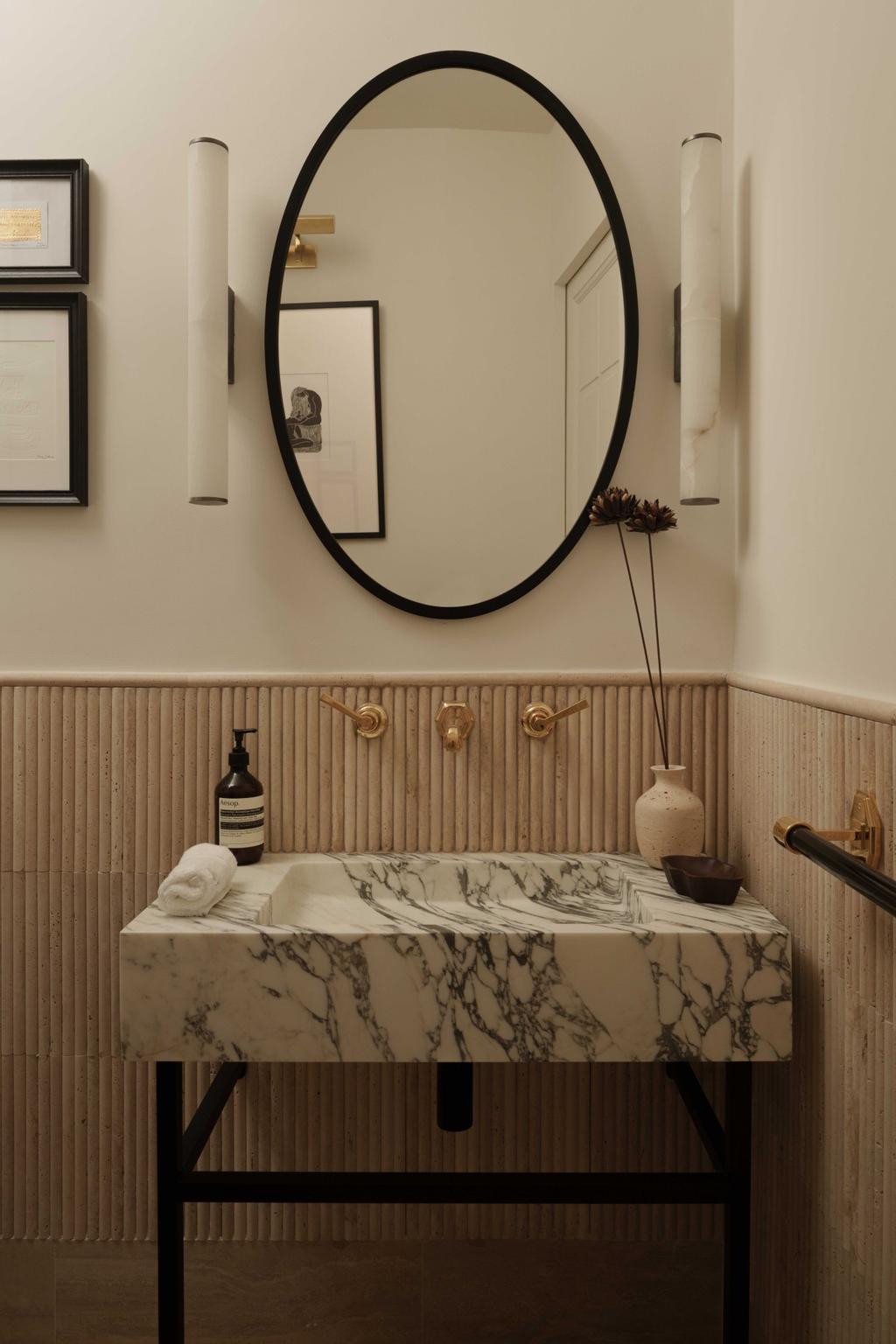 A refined Parisian powder room with a sculptural marble sink, vertical ribbed wall paneling, and a bold black-framed round mirror. Subtle gold fixtures and minimal decor elevate the space, showcasing the essence of Parisian elegance through natural stone and timeless design elements.