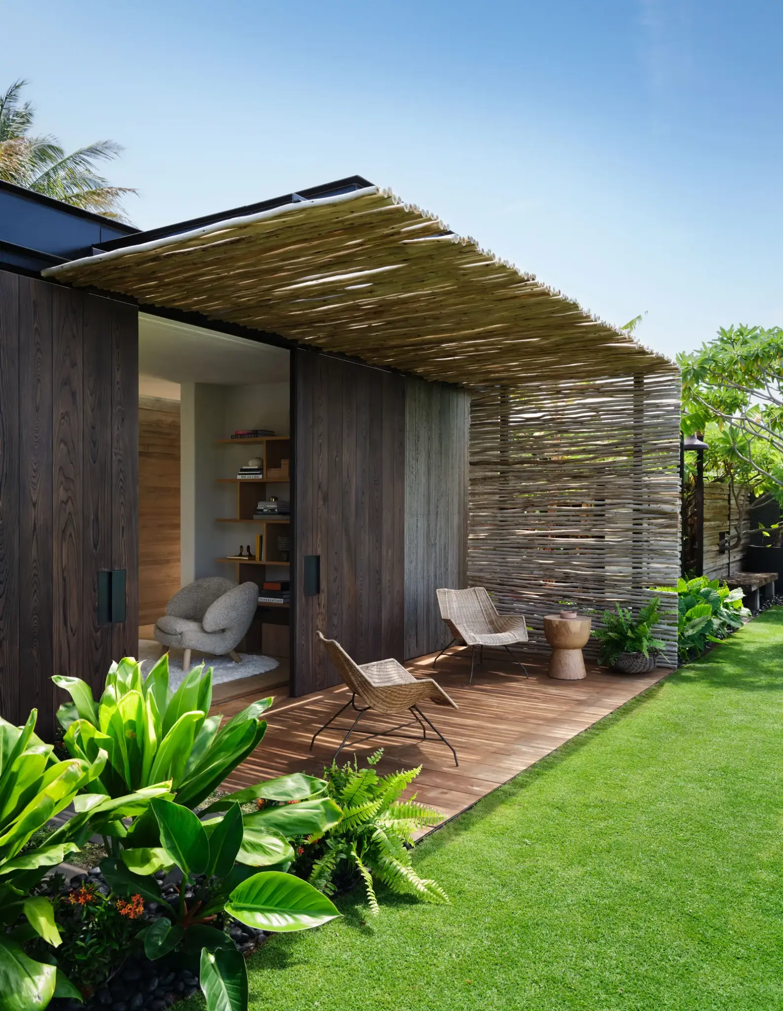 Tropical guesthouse exterior with lush green lawn, wooden pergola, and outdoor seating in a serene garden setting.

