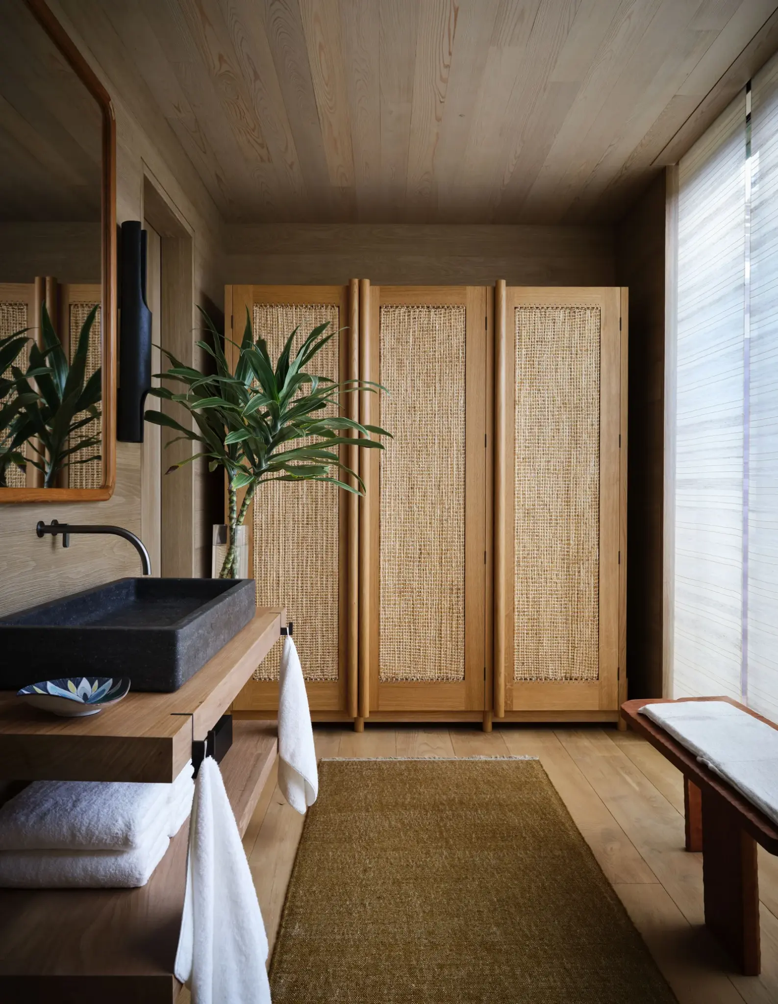 Minimalist tropical interior with natural wood finishes, woven cabinet doors, indoor plants, and soft lighting. A serene blend of textures and organic elements creates a warm and calming atmosphere.

