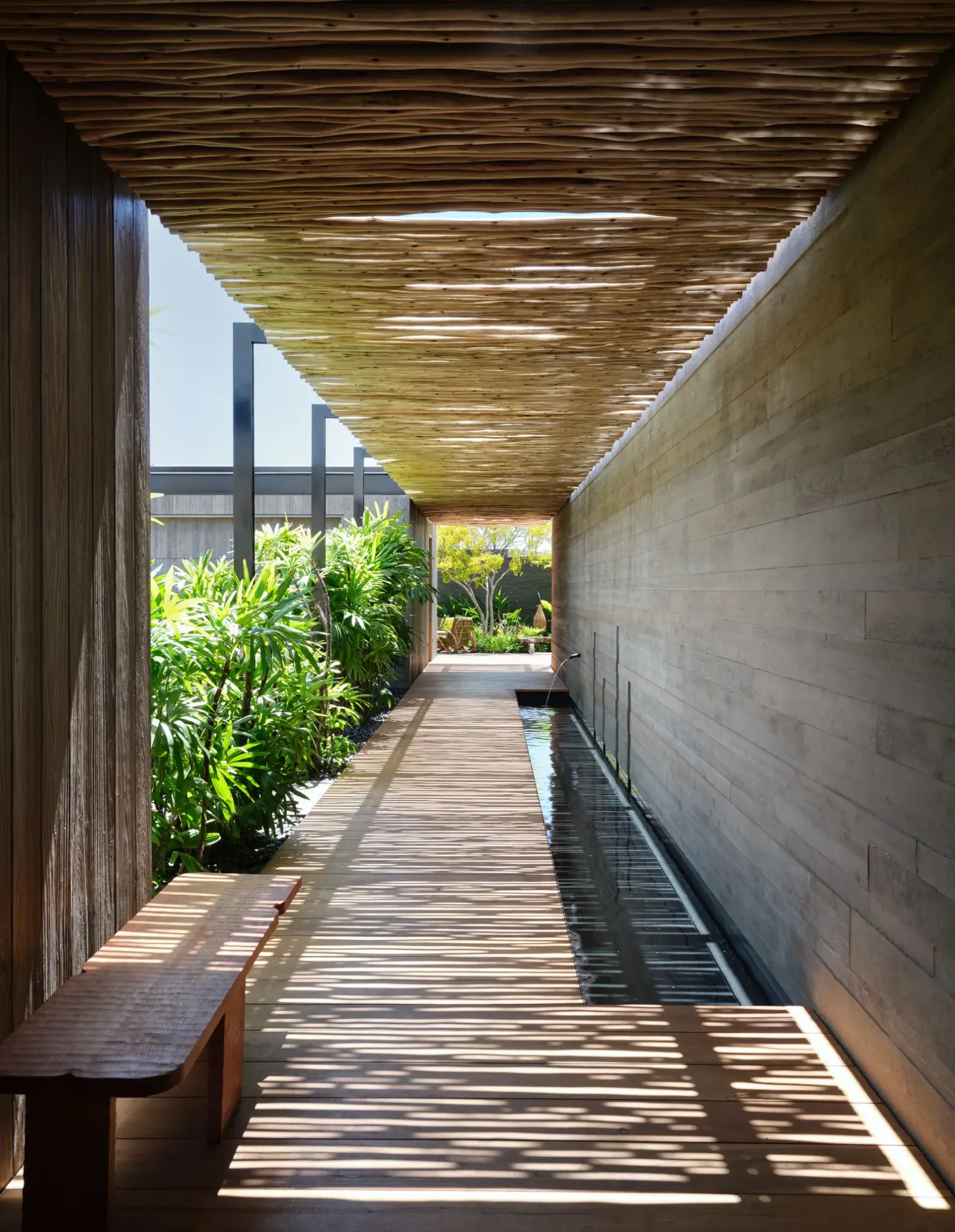 Open tropical hallway with wooden ceiling and lush green plants, leading to a bright garden area. Natural materials blend with the outdoor setting.

