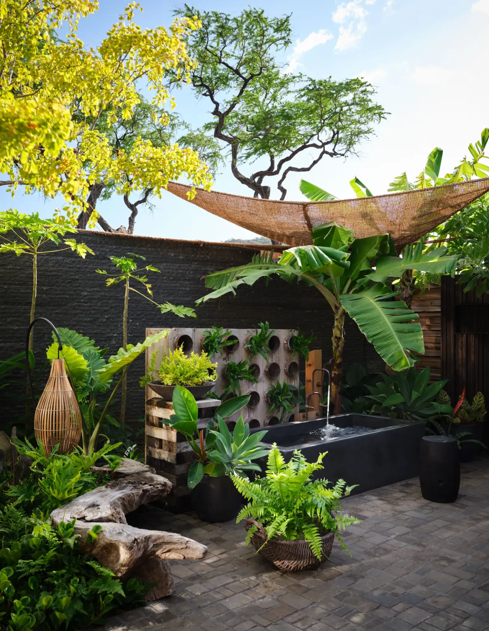 Lush tropical courtyard featuring a stone bathtub, bamboo fencing, and dense greenery under dappled sunlight. A peaceful, spa-like outdoor space designed for relaxation and connection with nature.

