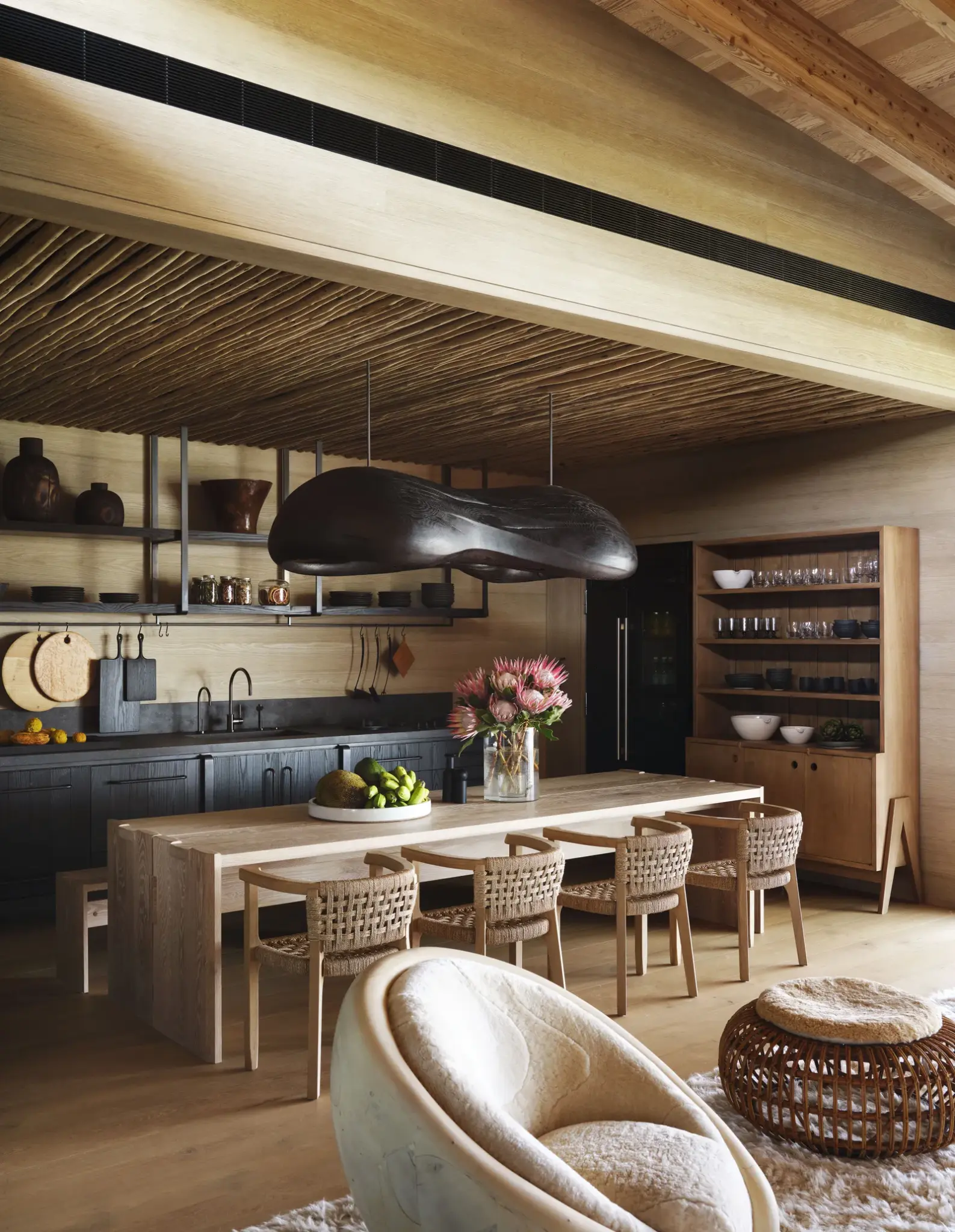 A stylish tropical kitchen and dining area featuring natural wood finishes, a woven ceiling, and modern pendant lighting. The design highlights organic materials, warm tones, and handcrafted furniture, creating a functional yet inviting space aligned with tropical house design aesthetics.

