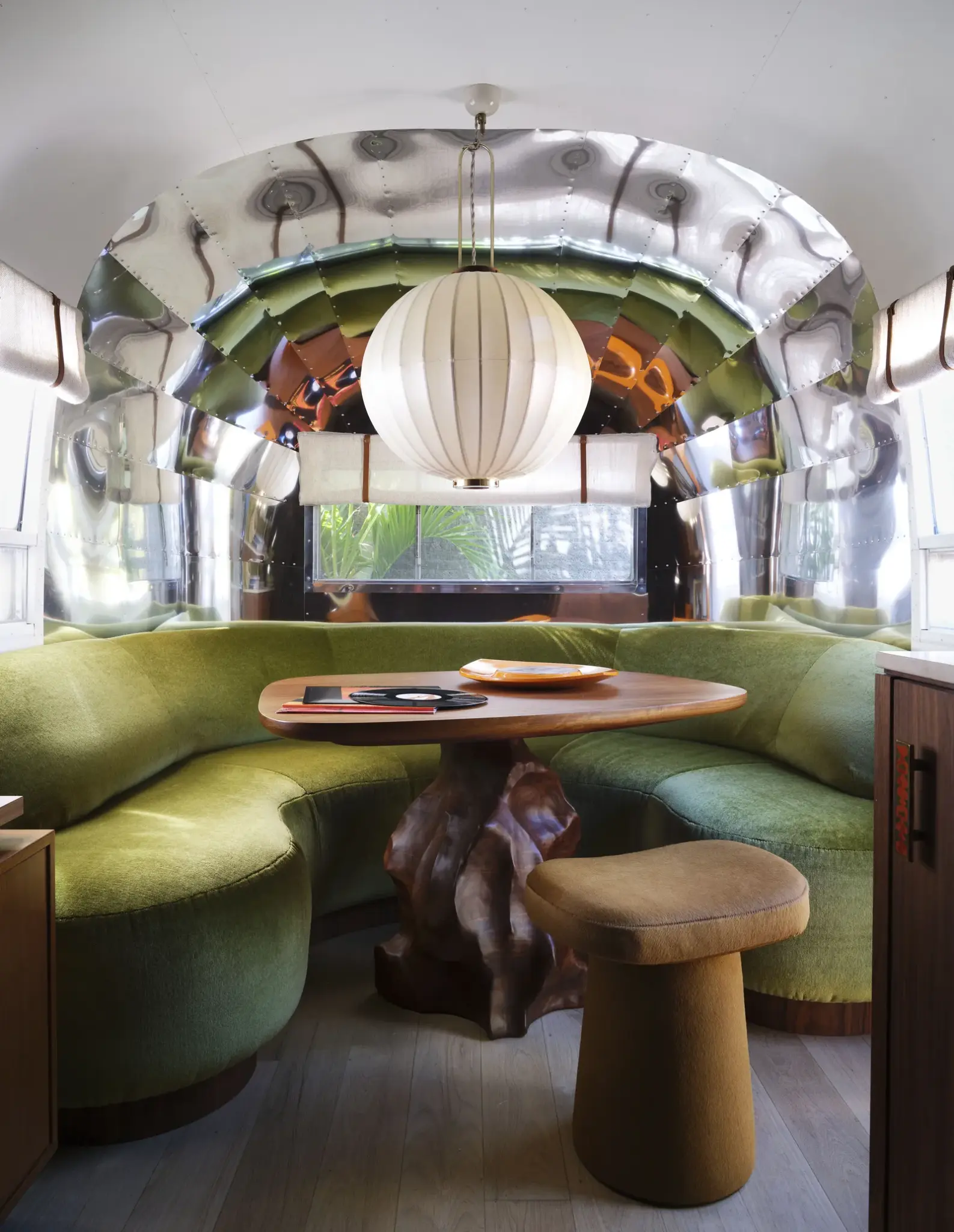 A unique tropical lounge area with curved green seating, a sculptural wood table, and a dramatic central pendant light. Surrounded by reflective surfaces and soft textures, this space blends retro charm with modern tropical design flair.

