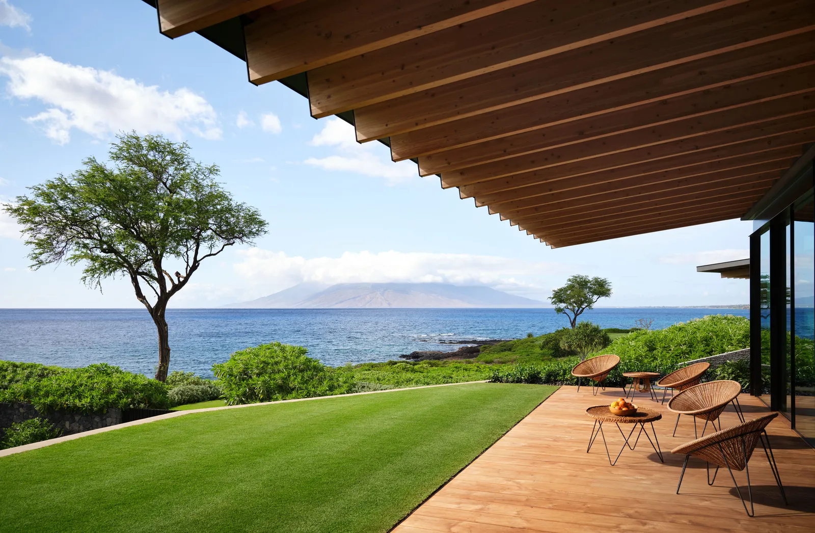 Modern tropical patio with wooden pergola, ocean view, and minimalistic outdoor furniture on a wooden deck. Lush green lawn and palm trees create a serene island atmosphere.