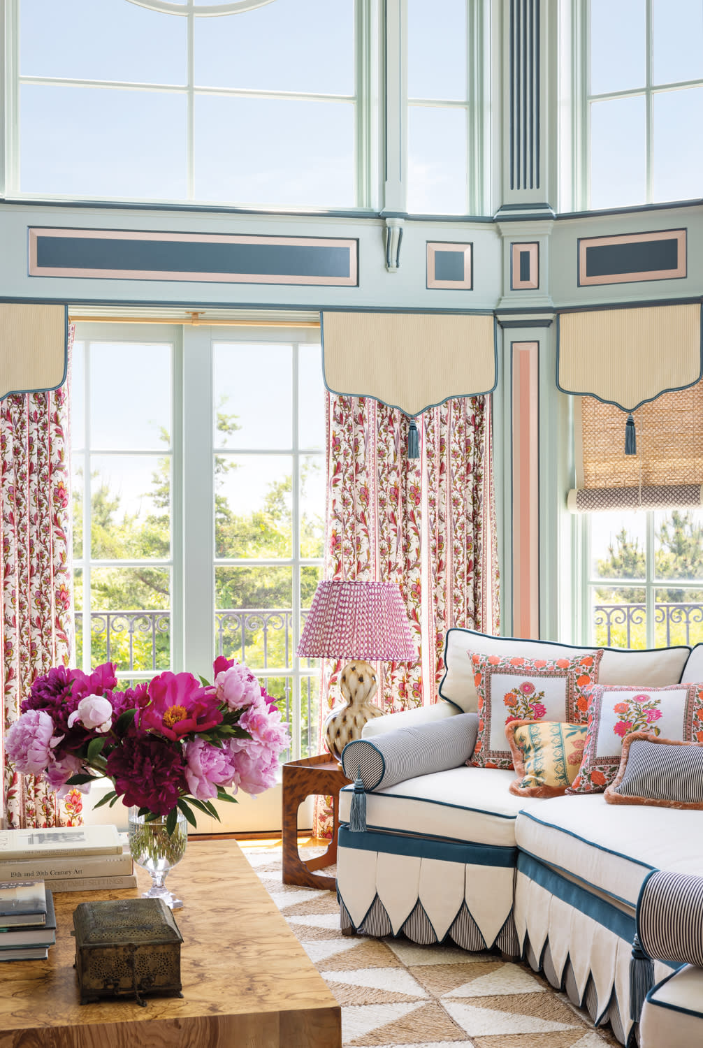 Bright living room with floral curtains, scalloped valances, and a tailored blue sofa accented with colorful embroidered pillows. A geometric rug and fresh flower arrangement complete the eclectic, elegant space.

