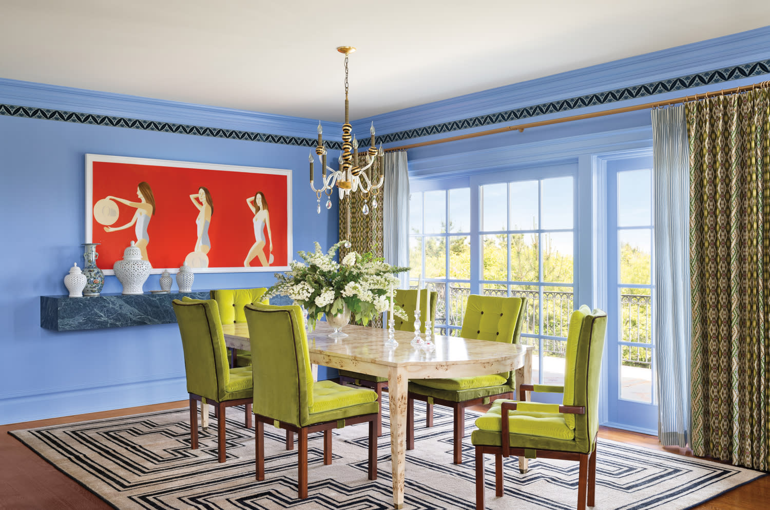 Eclectic dining room featuring a round marble table surrounded by vivid chartreuse velvet chairs. Blue-painted walls, a bold red artwork, and a classic chandelier add contrast and personality to the layered design.

