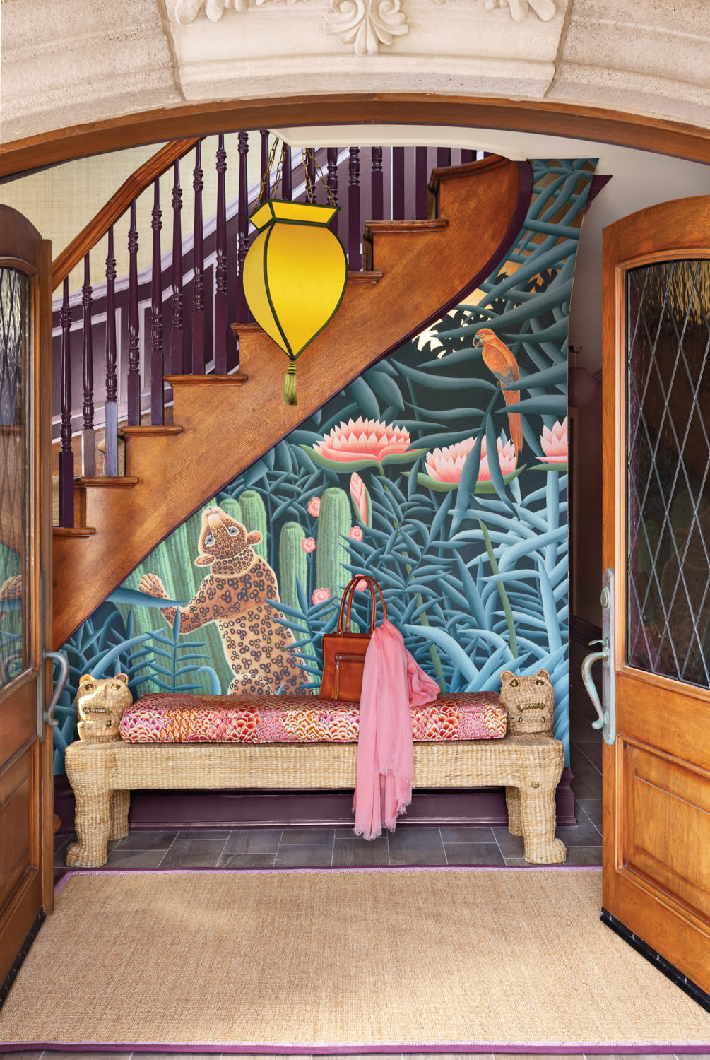 Vibrant home entryway with tropical jungle mural, wicker bench with animal-shaped arms, and colorful accessories under a wooden staircase.