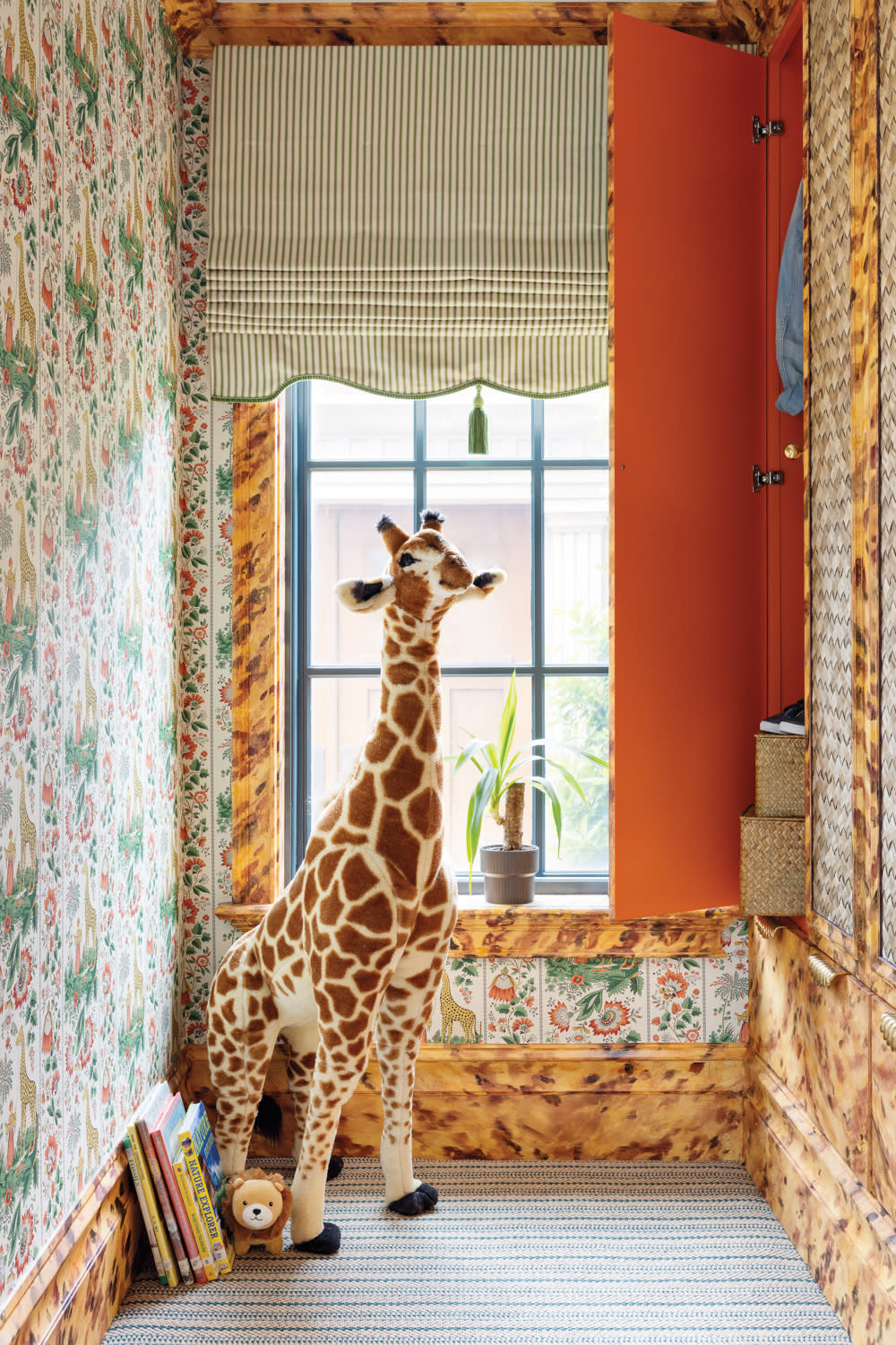Cozy reading nook with floral wallpaper, tortoiseshell paneling, and a large plush giraffe standing near a window. A low bookshelf with children’s books adds charm to the colorful, eclectic design.