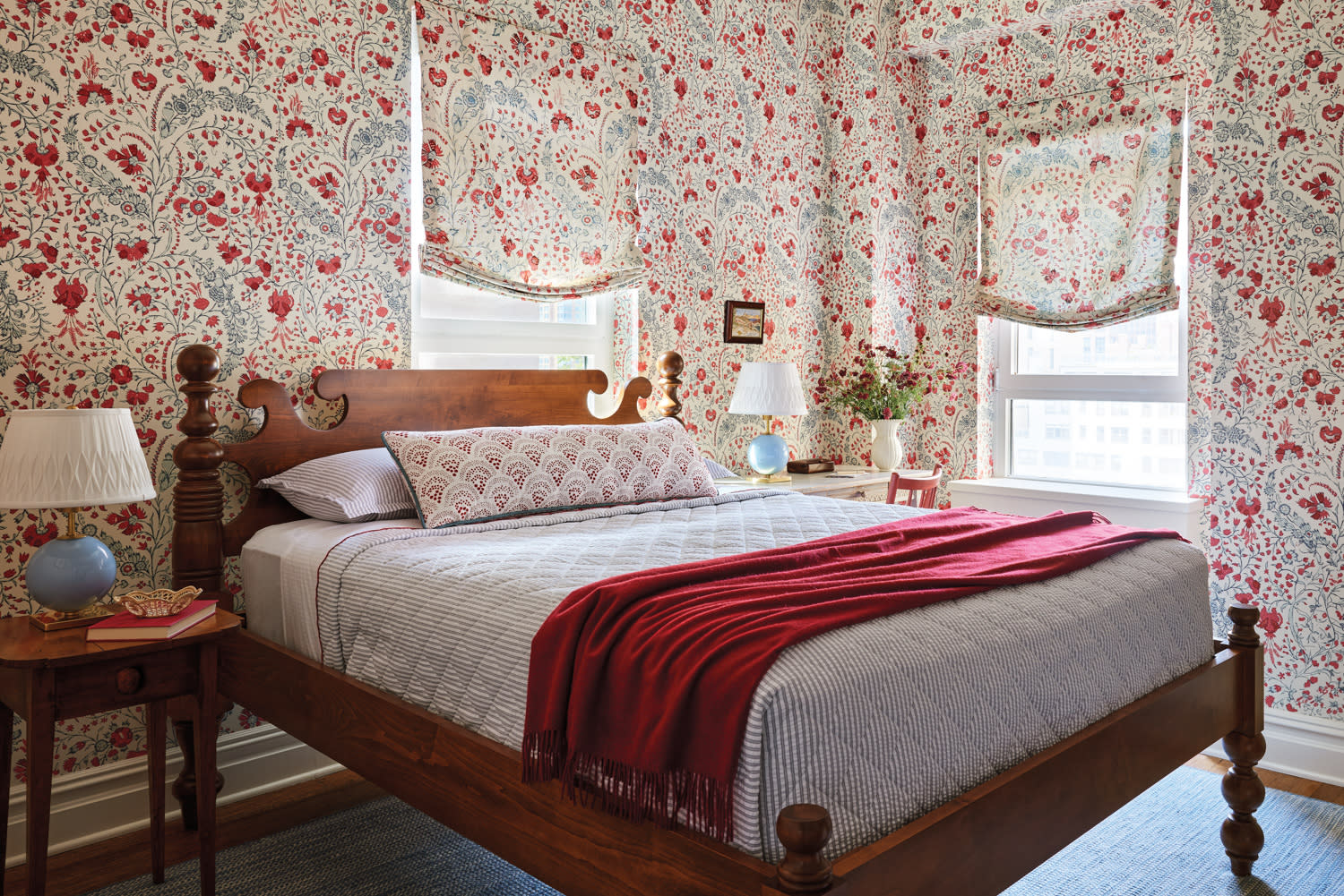 Cozy bedroom with floral wallpaper, wooden bed frame, red blanket accents, and a mix of traditional and eclectic decor elements.