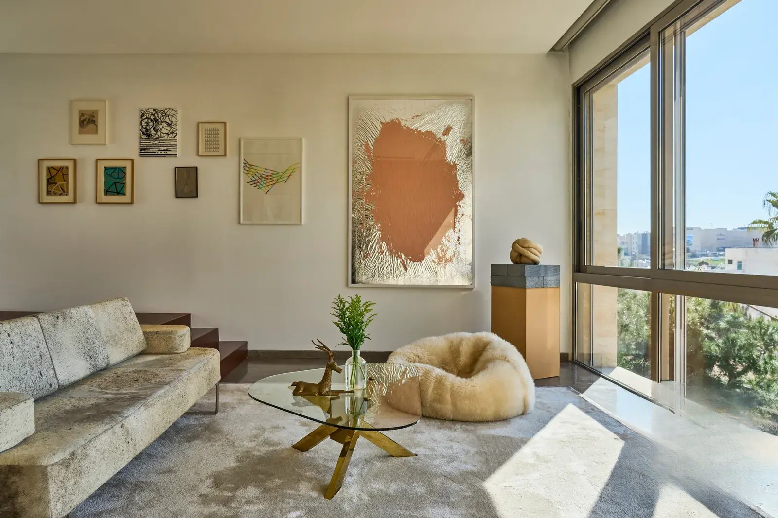 Large floor-to-ceiling window with views of greenery, a spacious area featuring a soft sofa, puff, and a glass round coffee table. Artworks on the walls and a unique handmade rug complete the space