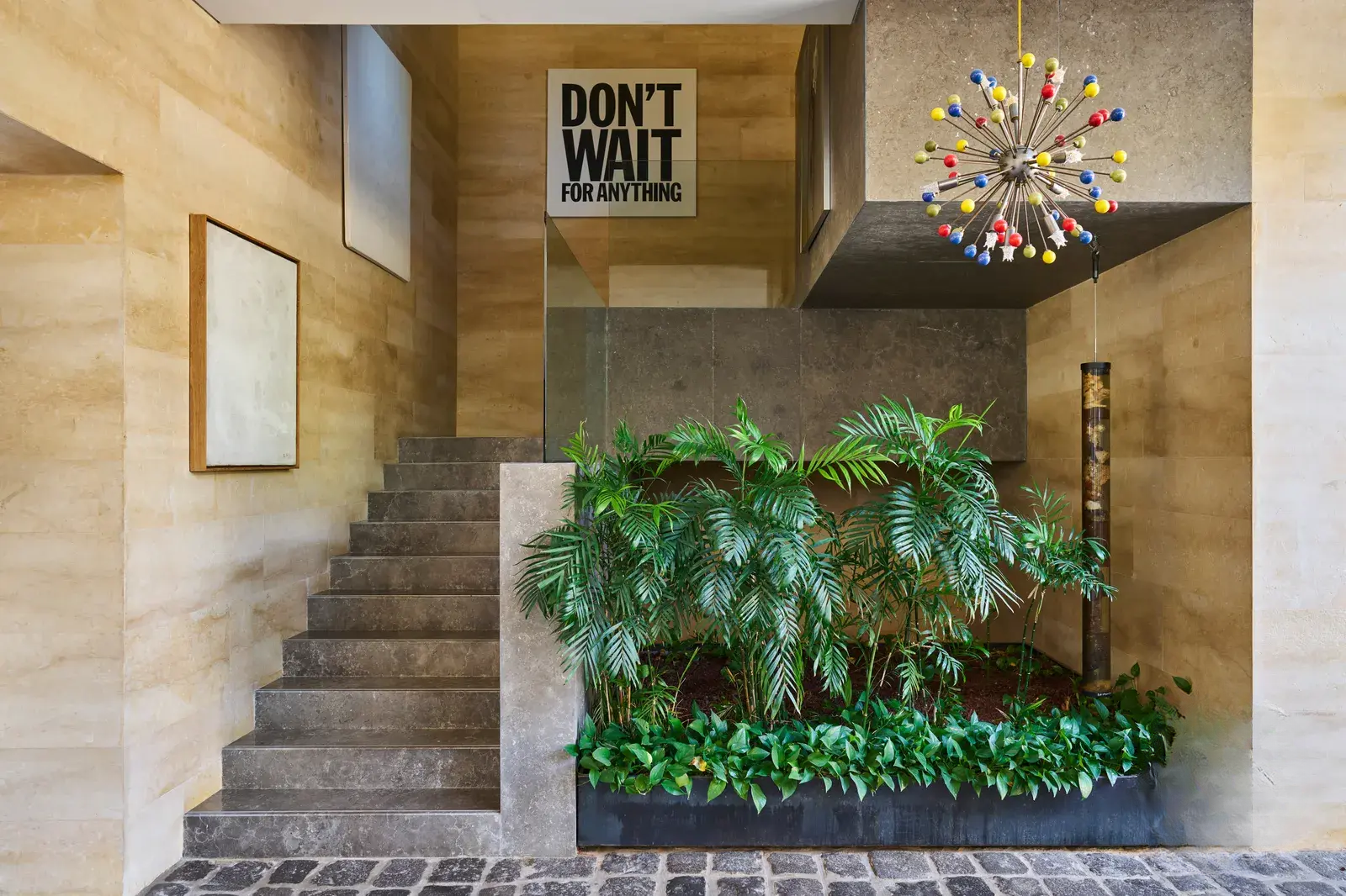 Staircase leading to the upper floor, surrounded by lush greenery, with Sputnik-style lighting, a stone pathway, and art pieces displayed on the walls.