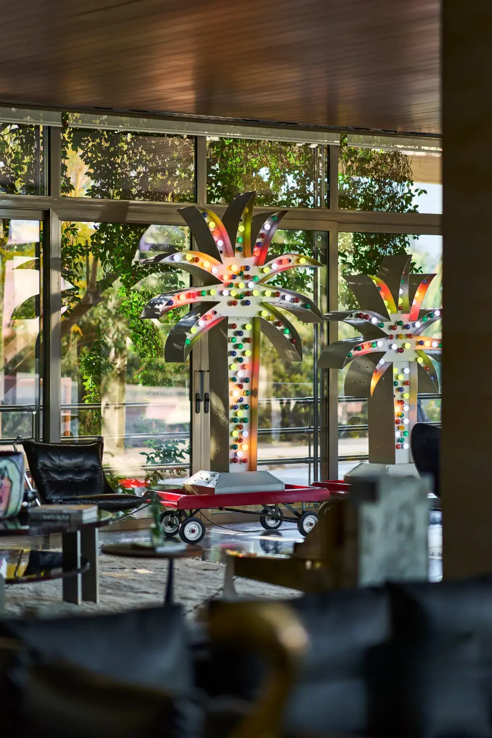 Bright and colorful electric-powered sculptures of palm trees on wheels, with a kid's toy car beside them, next to a large floor-to-ceiling window letting in abundant natural light in the living room