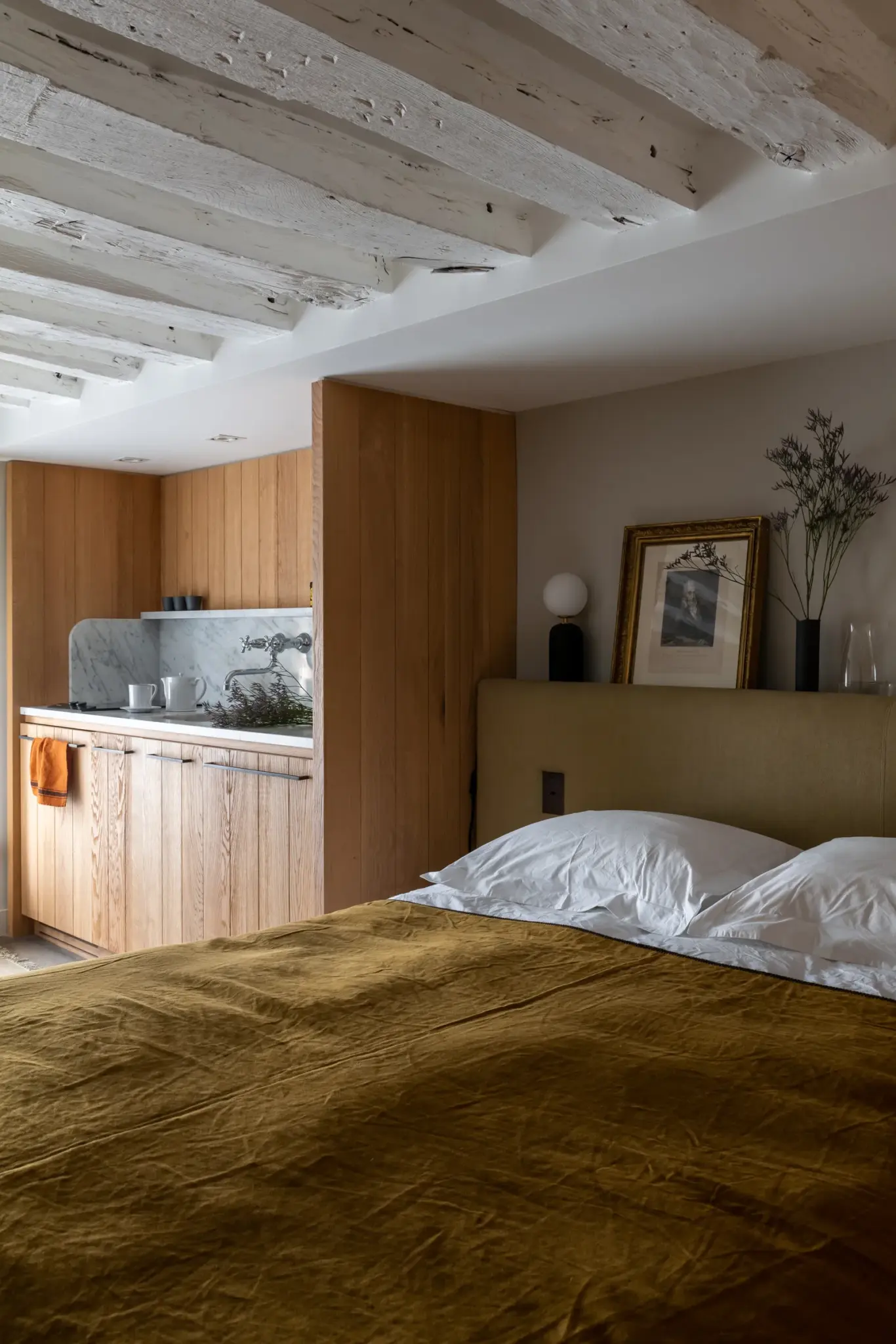 Double-size bed next to a kitchenette, accented with decorative lighting, art works, and natural colors for a cohesive and calming atmosphere.