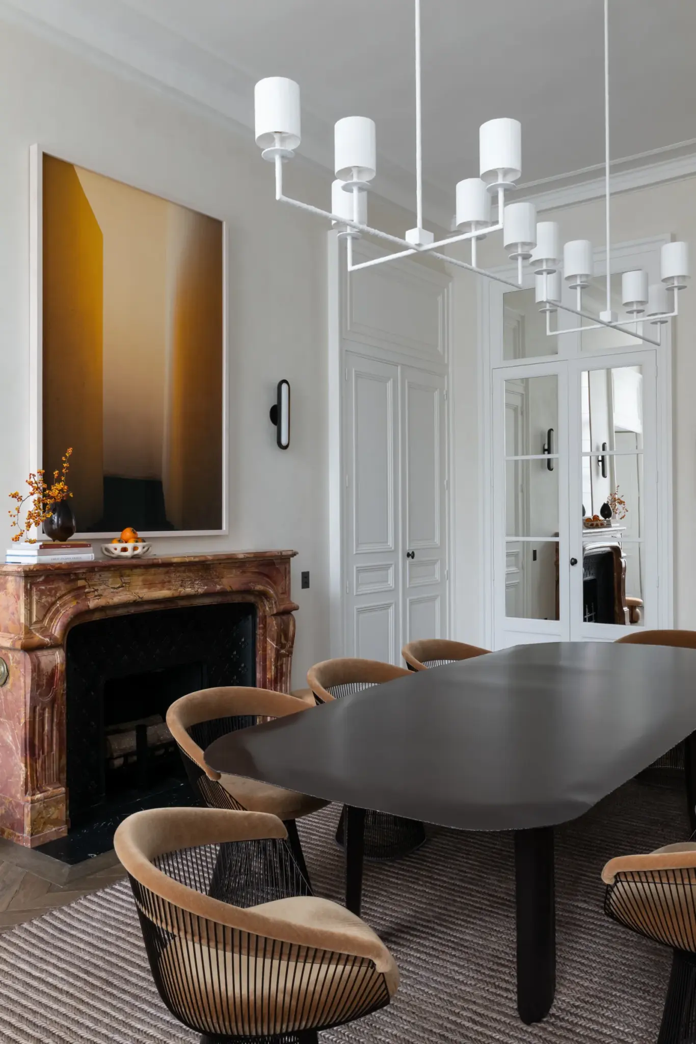 Dining area with a large table, handmade chairs, positioned next to a fireplace, complemented by designer lighting and plenty of natural light.