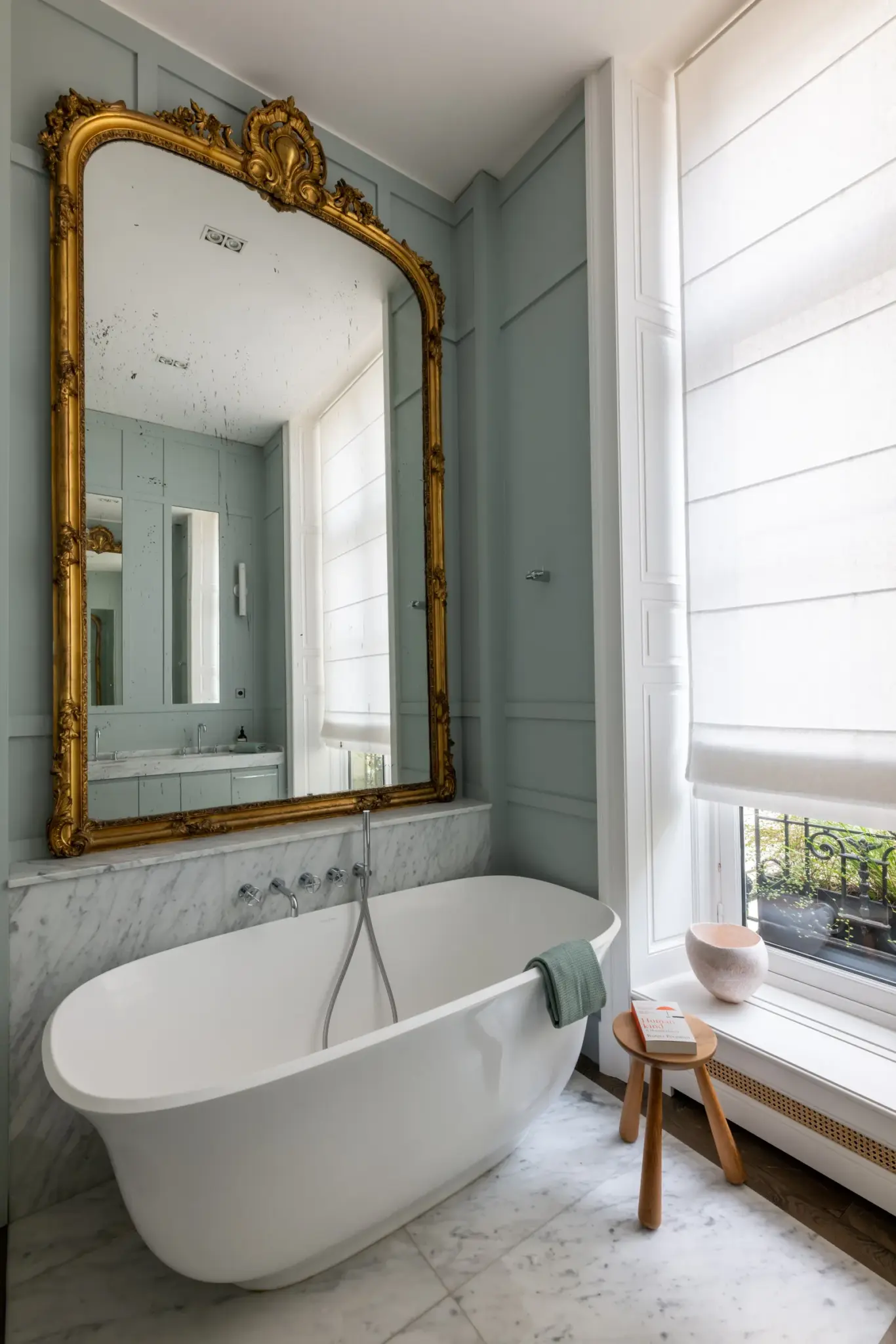 Bathtub in the studio paired with a handmade wooden chair, large metal-framed mirror, and a huge window letting in plenty of natural light