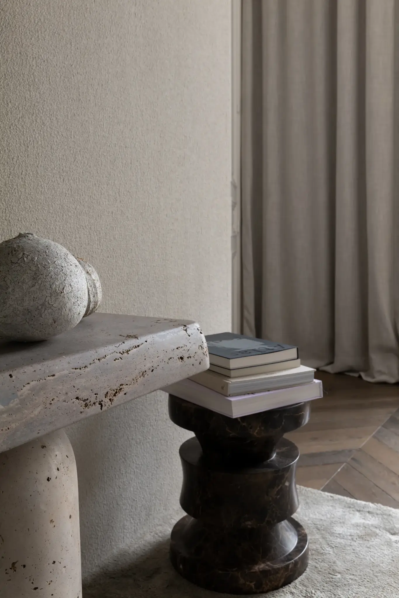 Books arranged on a small round wooden table next to a stone table, set on wooden parquet flooring and bathed in natural light.