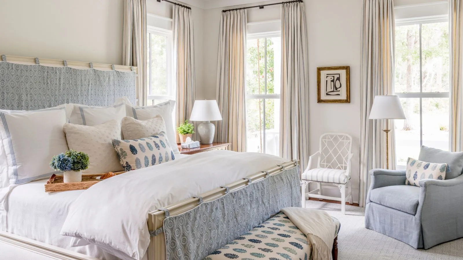 This luxury bedroom boasts a bright, airy feel with soft blue and white tones. Patterned fabrics adorn the bed and seating, while multiple windows bring in natural light, creating a serene and inviting space.