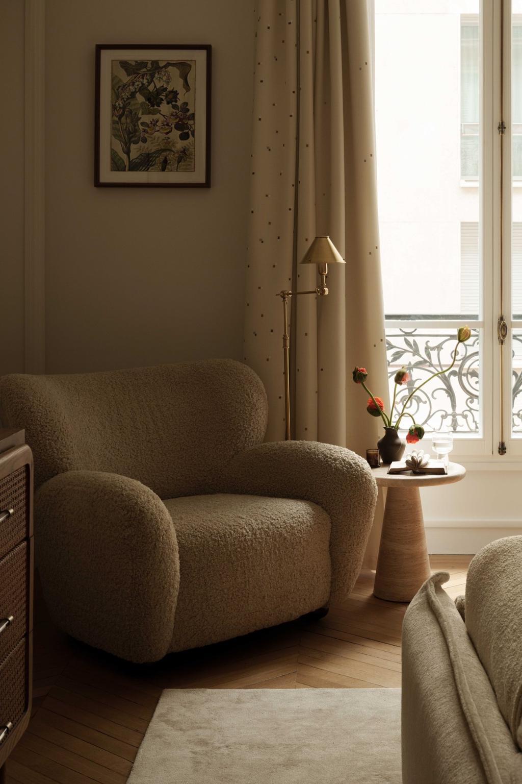 A cozy Parisian reading nook with a boucle armchair, round wooden side table, and brass floor lamp. Set against tall French windows with flowing curtains and delicate floral decor, this corner combines comfort and elegance in true Parisian interior design fashion.