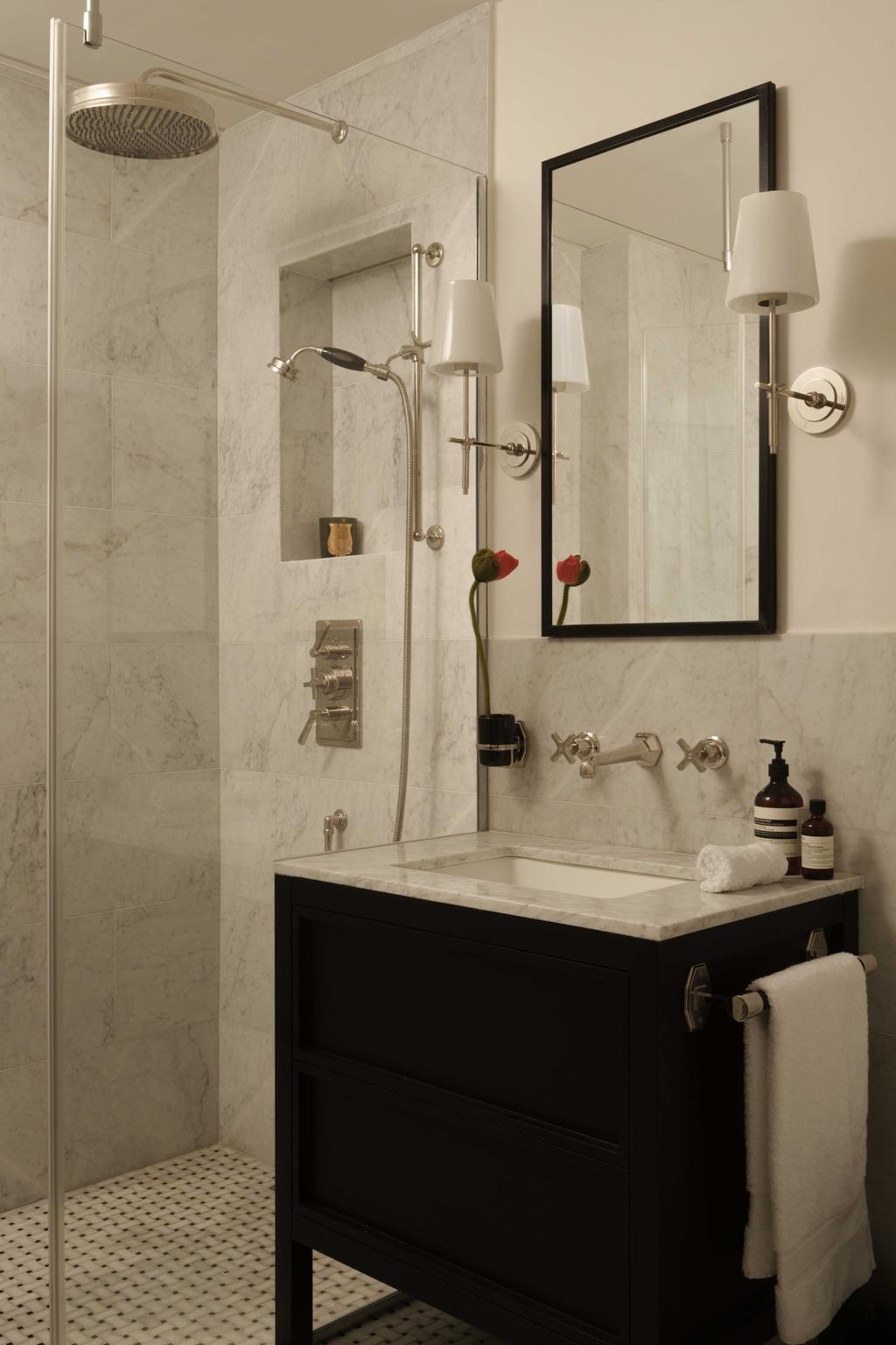 A sophisticated Parisian bathroom featuring a walk-in marble shower, black vanity with dual mirrors, and elegant wall-mounted lighting. Brass accents and red floral touches add warmth to the classic interior, combining modern luxury with Parisian charm.