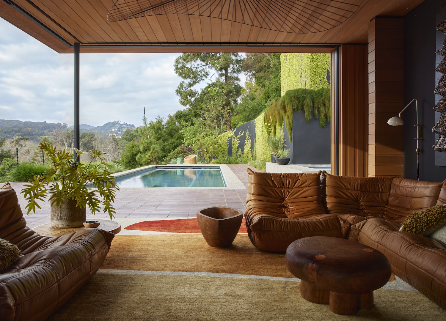 A cozy living room with plush leather seating and a view of a pool. Warm wood ceilings and earthy tones capture the 70s interior design aesthetic.
