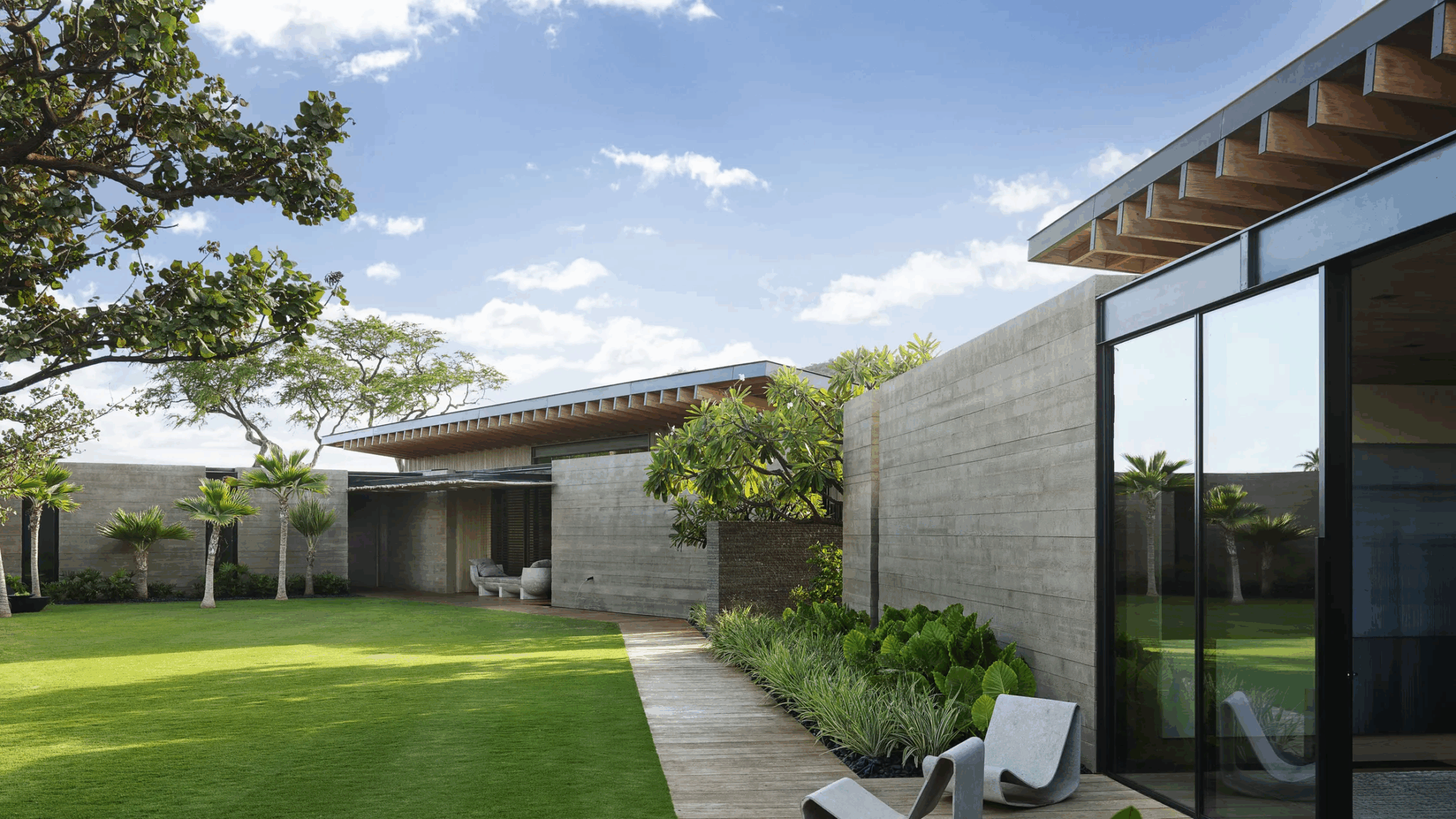 A stunning tropical house exterior with modern concrete architecture, lush green landscaping, and expansive glass windows. The design blends indoor and outdoor living, showcasing a serene courtyard with minimalist lines and natural materials under a bright blue sky. Perfect inspiration for tropical house design ideas.

