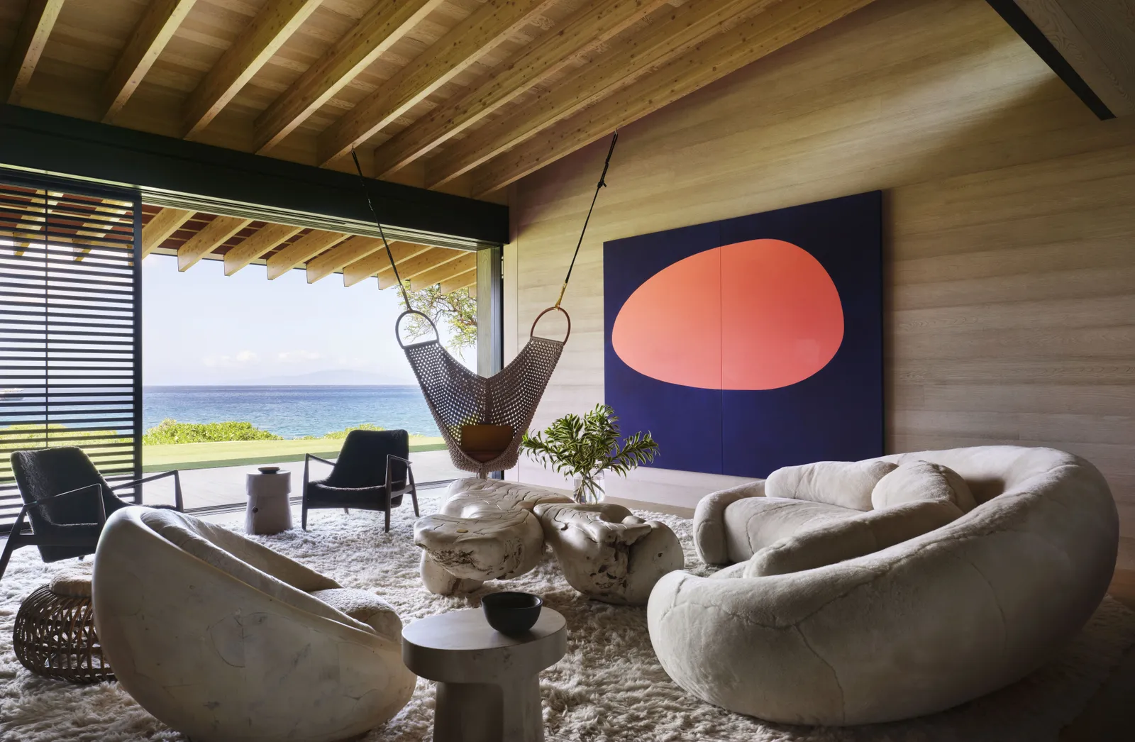 A cozy tropical interior with ocean views, featuring plush curved furniture, a hanging hammock chair, and a bold abstract wall art piece. Exposed wooden beams and natural textures create a relaxing indoor-outdoor flow, ideal for tropical house design lovers seeking comfort and style.

