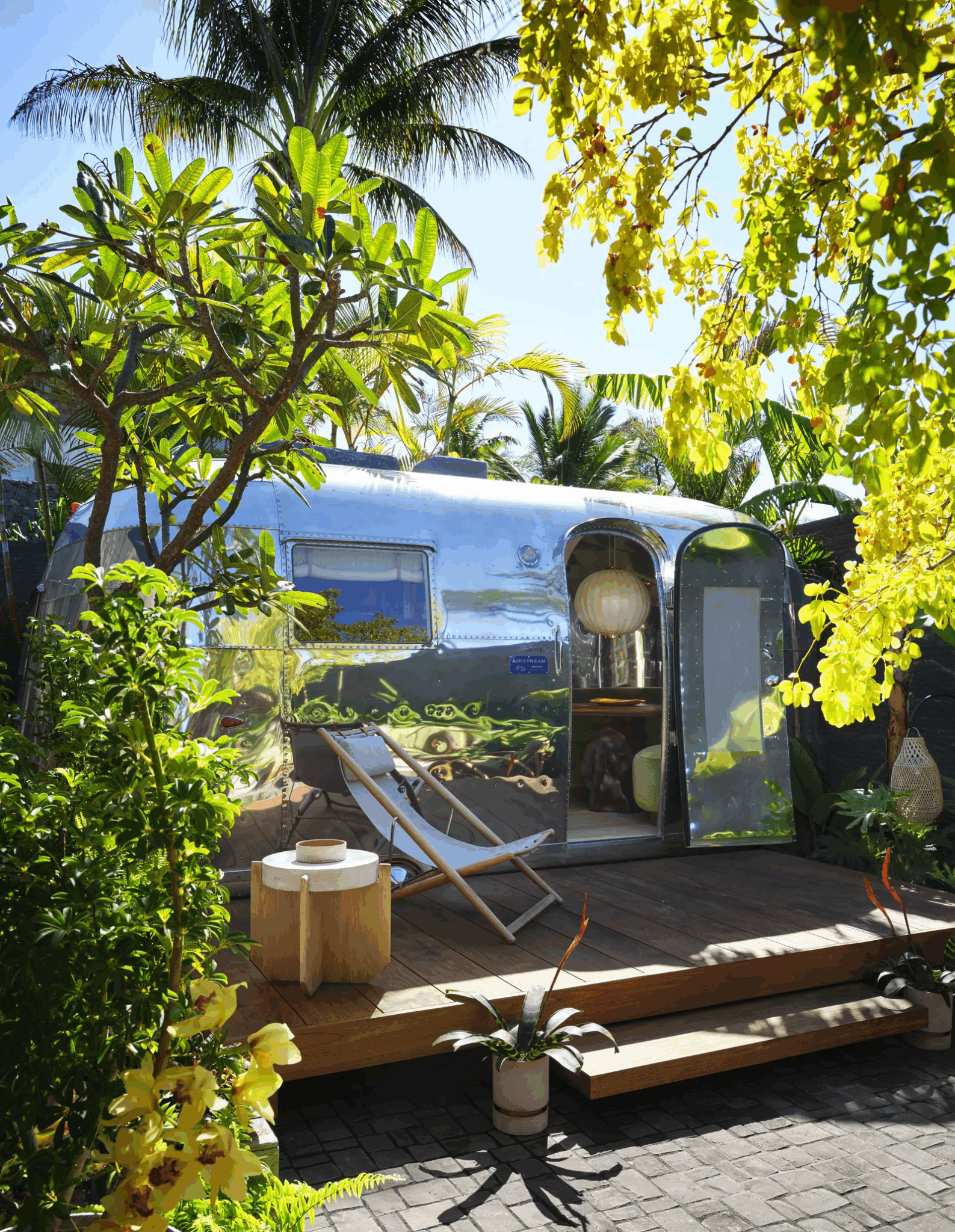 Stylish Airstream trailer surrounded by lush tropical plants and trees, with wooden deck and modern outdoor furniture. Sunlight filters through the leaves, creating a cozy and vibrant atmosphere.

