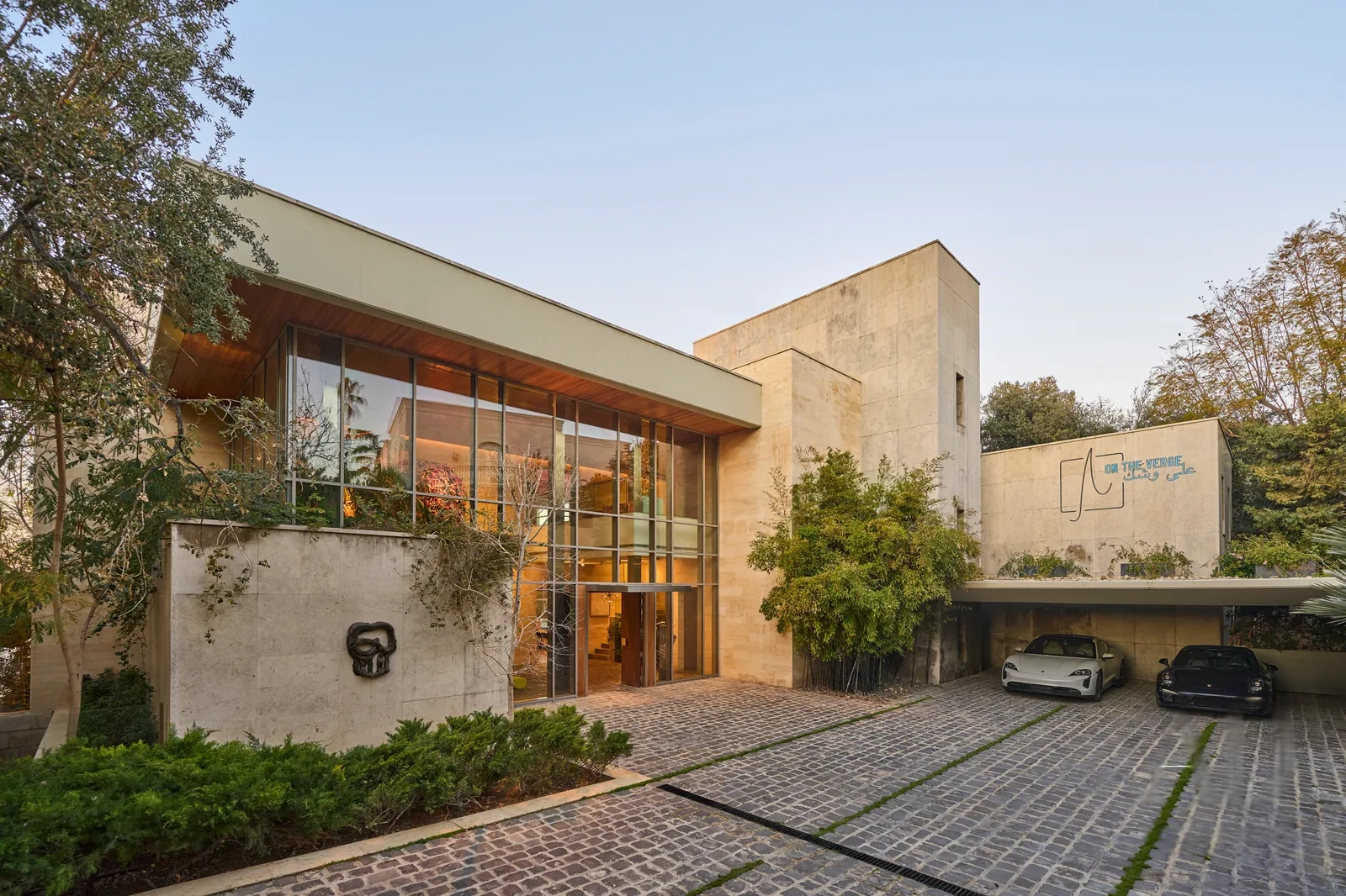 Exterior view of the villa with a cobblestone road, luxury cars parked in the driveway, large floor-to-ceiling windows, and surrounded by lush nature